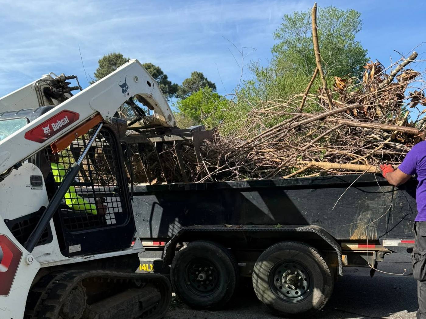 Skid steer loading cut brush and tree limbs into a black dump trailer during a property clearing and hauling job